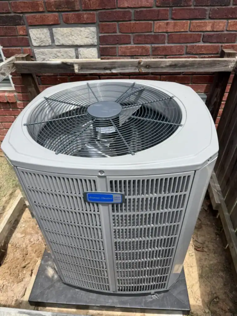 New American Standard air conditioner on a black pad beside a red brick wall